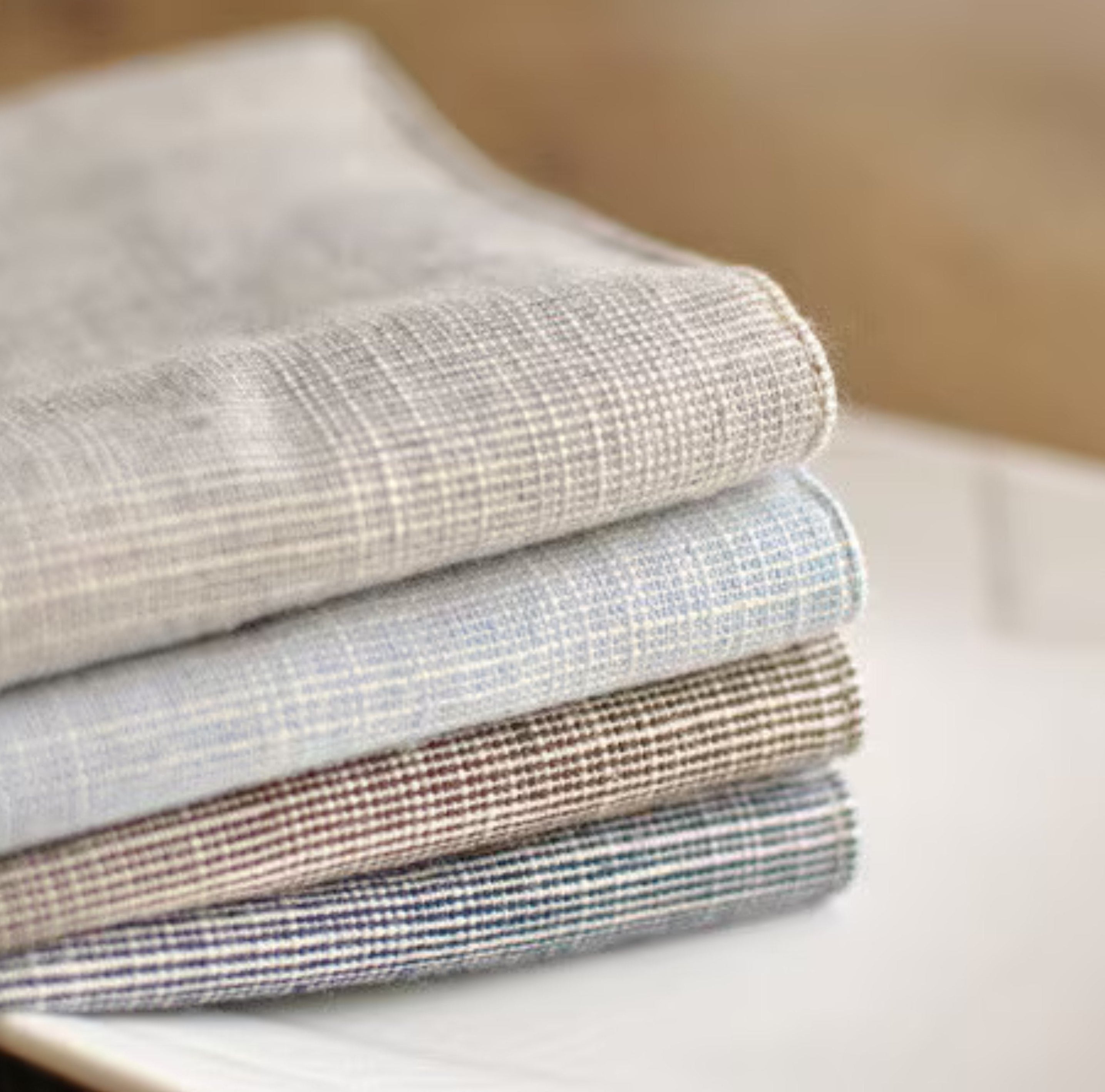 Four folded woven fabric napkins or cloths stacked on a white surface, shown in soft neutral tones with subtle plaid textures.
